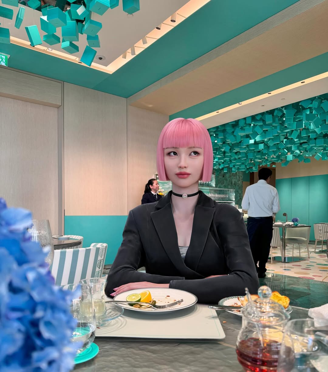 Virtual influencer Imma with pink bob haircut in black blazer sitting at upscale turquoise restaurant with food plate and cocktail