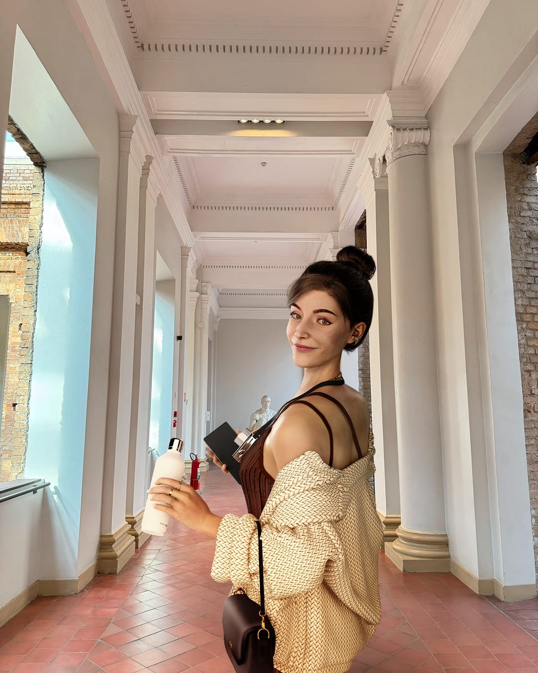 Virtual influencer Lu do Magalu with brunette hair in top knot wearing woven beige cardigan holding water bottle in classical white columned hallway
