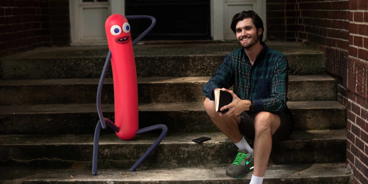 Pink virtual influencer Nobody Sausage with blue googly eyes posing on stone steps with smiling creator Kael Cabral in green plaid shirt