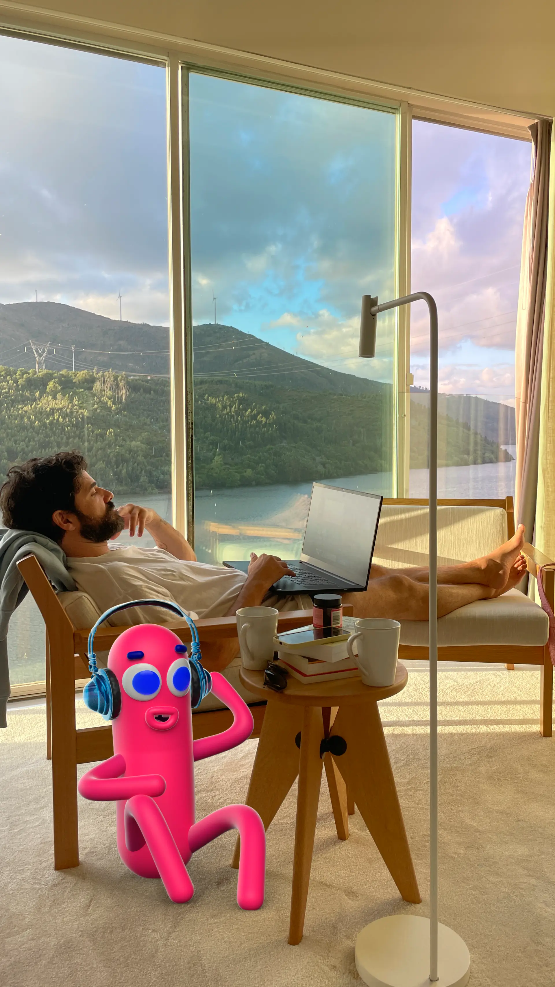 Virtual influencer Nobody Sausage with pink cylindrical body and headphones sitting beside creator Kael Cabral working on laptop in modern apartment with mountain lake view
