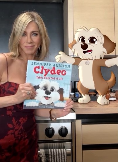 Virtual influencer Clydeo animated puppy character with Jennifer Aniston holding children's book titled Clydeo Takes a Bite Out of Life in modern kitchen setting